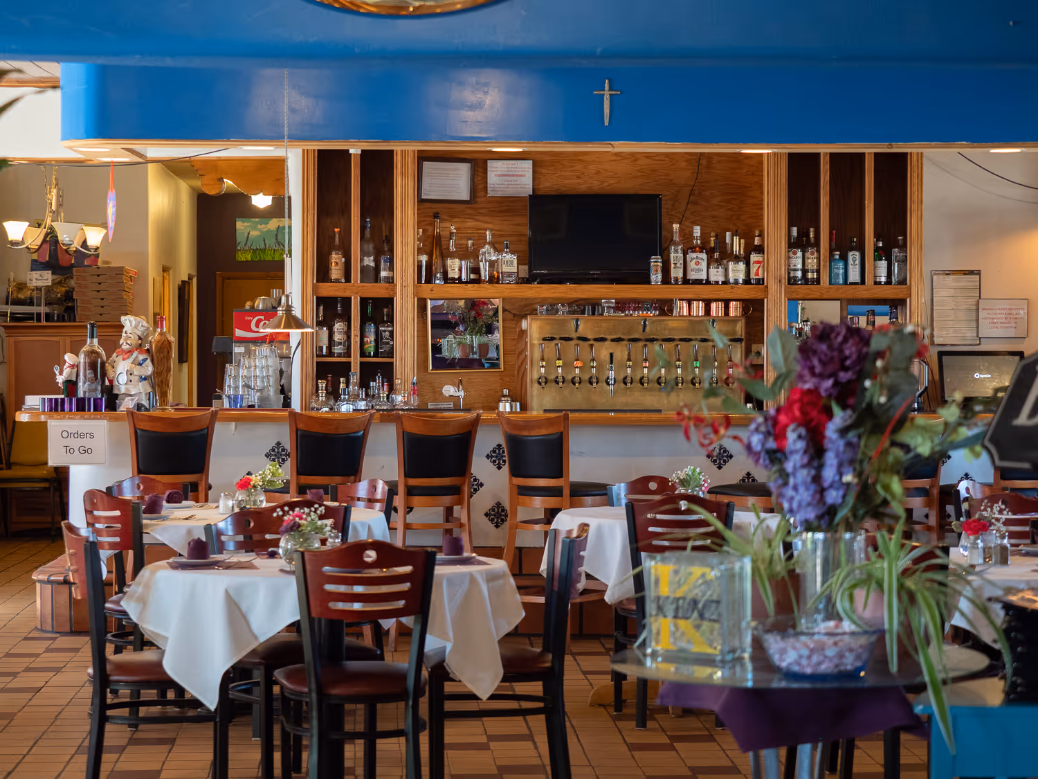 The bar with stools, shelves of bottles and beer taps, behind set dining tables and fresh flowers