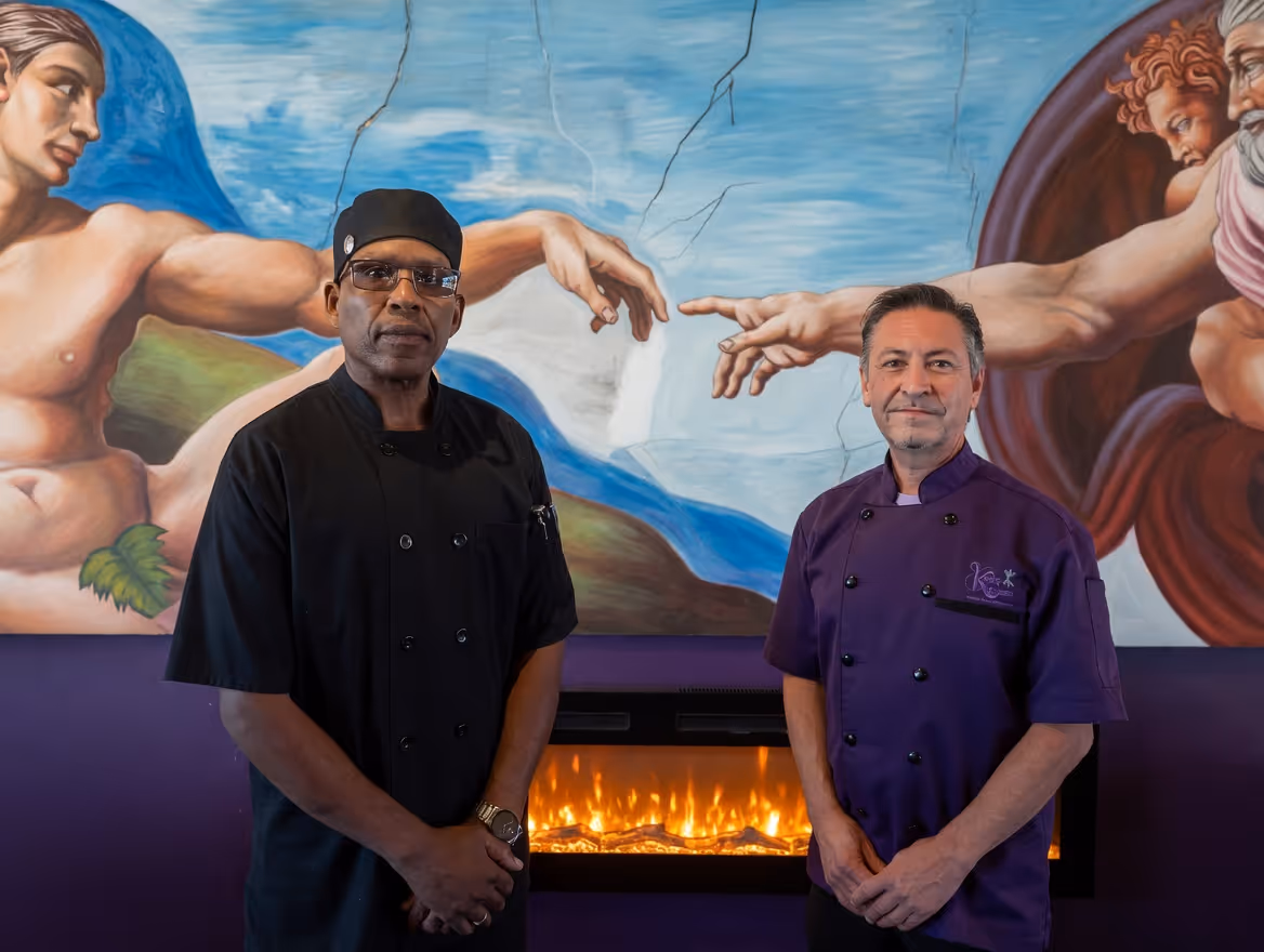 The two chefs in chef's coats in front of a Creation of Adam mural and a fireplace
